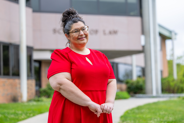 Mid shot of Professor Dyszlewski wearing a red dress with hands clasped in front, hair in an updo bun, wearing eyeglasses with School of Law in the background.