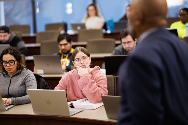 A law student in a pink sweater listens attentively to a professor during class, with other students working on laptops in the background.