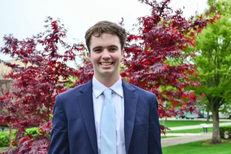 Medium shot of subject Nate Reid wearing a jacket and tie, smiling, with fall colored leaves in the background.