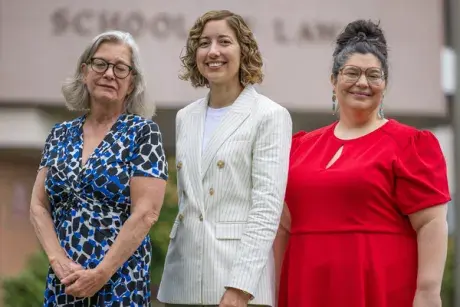 Three women faculty members--left wearing floral blue dress with glasses, middle wearing an off white suit jacket, right wearing a bright red dress and eyeglasses with the School of Law building sign visible in the background.
